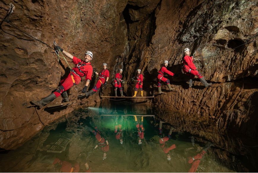 6 people in red jumpsuits, holding onto ropes in a cave